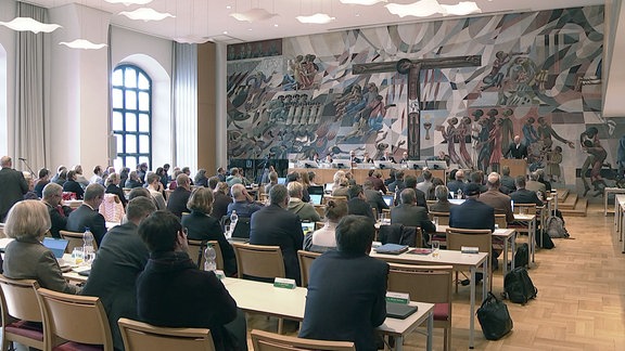 Menschen sitzen in einem großen Saal mit einem Wandgemälde an ihren Tischen.