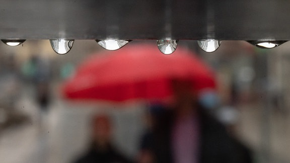 Symbolbild: Regentropfen hängen im Vordergrund, im Hintergrund unscharf ein roter Regenschirm.