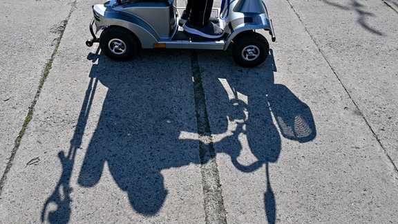 Der Schatten eines Rentners, der mit einem Elektromobil, auch E-Quad oder Seniorenmobil, unterwegs ist, ist auf dem Weg zu sehen.