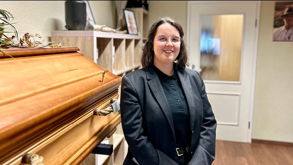 Bestatter-Azubi Franziska Ritscher trägt einen dunklen Blazer und eine schwarze Bluse. Sie steht im Ausstellungsraum vor einem hellbraunen Sarg aus Holz und schaut in die Kamera.