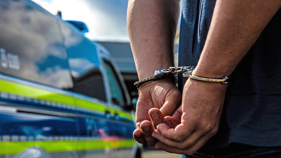 Eine Person wird durch die Polizei festgenommen, es wurden die Handschellen angelegt im Hintergrund sieht man den Streifenwagen der Polizei stehen. 