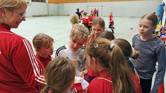 Mädchen in Sportklamotten stehen um eine lachende Frau in einer Turnhalle herum.