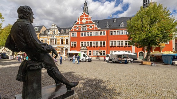 Bachdenkmal, ein junger Mann sitzt mit ausgestreckten Beinen auf einem Sockel, gegenüber ein altes Haus