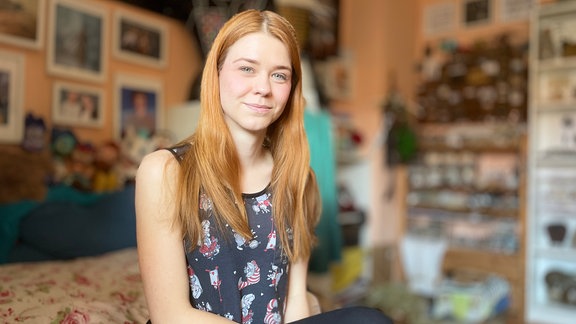 Eine junge Frau mit roten langen Haaren sitzt in einem Zimmer. Sie trägt ein weites Top und eine schwarze Strumpfhose. An den Wänden hängen Poster oder stehen Regale.