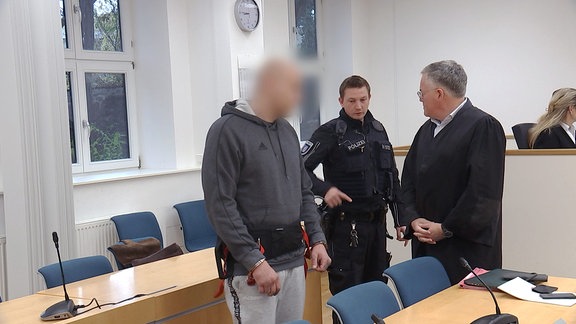 Ein Mann in Handschellen steht in einem Gerichtssaal neben einem Polizisten und einem Anwalt.