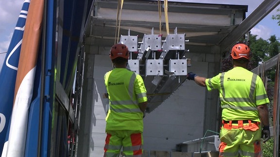 Zwei Handwerker haben ihre Hände an Stahlträgern, die an einem Kran hängen.