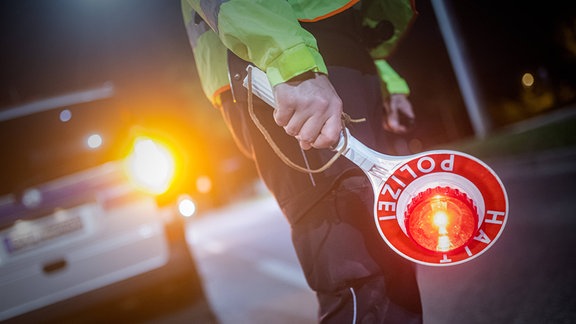 Eine Person mit Signalkelle der Polizei, im Hintergrund ein Einsatzwagen.