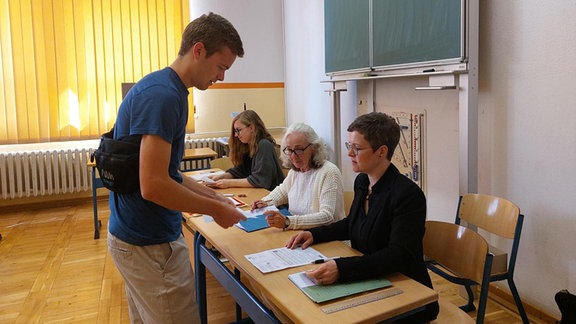 Ein Mann steht in einem Klassenzimmer vor einem Tisch, an dem drei Damen sitzen. Sie kontrollieren seine Personalien vor der Stimmabgabe. (Wahllokal in der Andreas-Gordon-Schule)