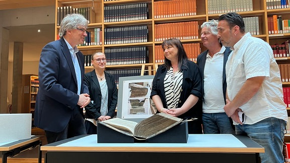 Fünf Menschen stehen in einer Bibliothek um ein altes Buch, das auf einem Tisch liegt.