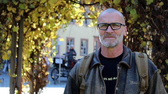 Ein Mann mit Zopf und Brille steht unter einem bunten Blätterdach auf der Straße in Weimar.