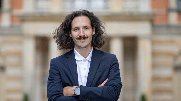 Der österreichische Regisseur Valentin Schwarz, ein Mann mit kinnlangen dunklen Haaren und Schnurrbart, steht mit verschränkten Armen vor dem Festspielhaus.