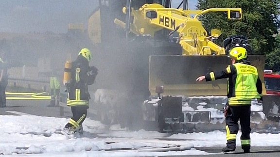 Eine Baumaschine auf einem Anhänger raucht stark. EIn Feuerwehrmann mit Atemschutz steht direkt an dem Fahrzeug und weiterer mit Abstand.