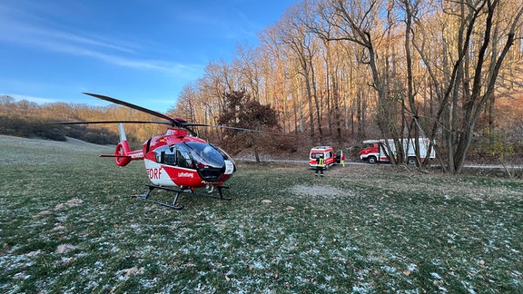 Ein Hubschrauber steht auf einer Wiese an einem Wald, dahinter ein Rettungswagen und ein Feuerwehrauto.