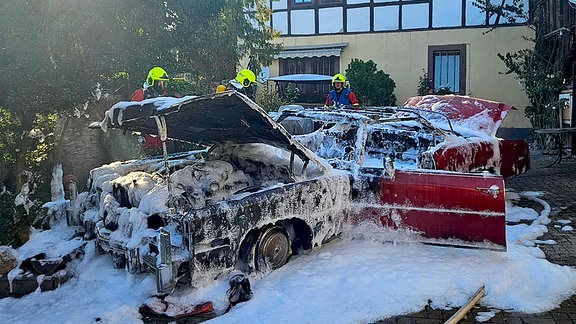 Ein ausgbranntes Auto in einer Einfahrt. Drei Feuerwehrleute stehen um das Fahrzeug. Viel Löschschaum.