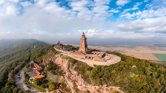 Luftaufnahme einer burgähnlichen Anlage mit hohem Turm auf einem bewaldeten Berg