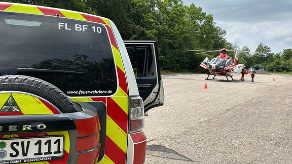 Ein Hubschrauber steht auf einer Betonfläche. Links ist ein Fahrzeug der Feuerwehr zu sehen. 