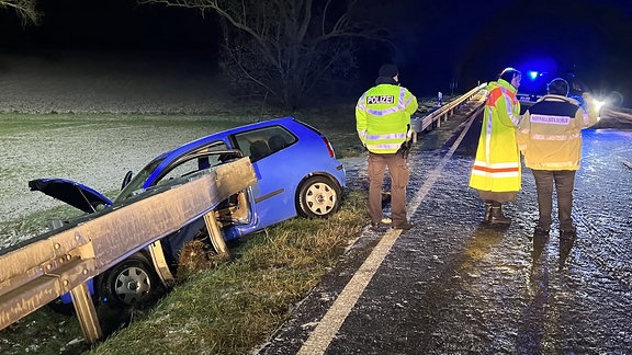 Ein blaues Auto steckt in der Leitplanke. Rettungskräfte begutachten den Unfallort.