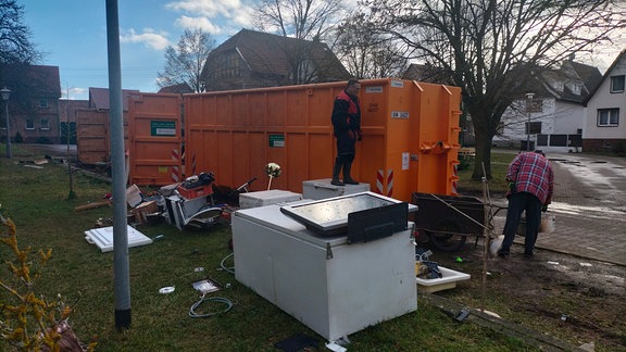 Zwei Männer räumen Hausratsgegenstände in einen Müllcontainer.