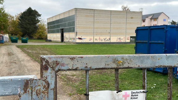 Ein Zauntor mit einem Schild des DRK mit der Aufschrift "Unbefugten ist der Zutritt verboten". Im Hintergrund eine alte Turnhalle in der das Katastrophenschutzzentrum des Deutschen Roten Kreuzes entstehen soll.