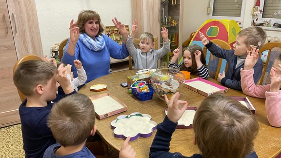 Eine Frau und mehrere Kinder sitzen an einem Tisch und strecken die Arme nach oben