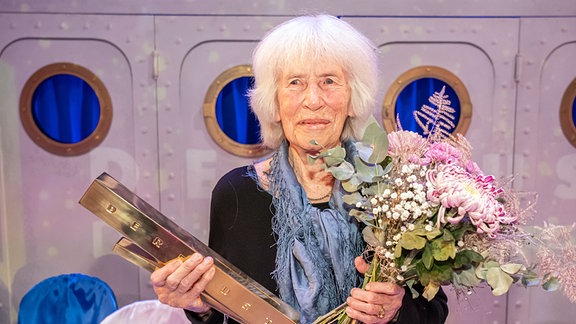 Nele Hertling, eine Frau steht auf einer Bühne und hält einen Preis und einen Blumenstrauß in den Händen.