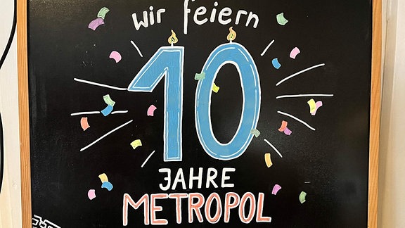 Auf einer bunt beschriebenen Tafel steht: "Wir feiern 10 Jahre Metropol"