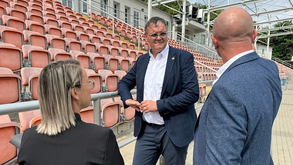 3 Personen vor der Tribühne eines Stadions