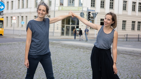 Zwei Frauen mit braunen Haaren und grau gemusterten Oberteilen lächeln in die Kamera. Ihre Hände berühren sich in einer zeigenden Geste nach hinten, die auf ein Gebäude verweist – die Tonhalle in Gera.