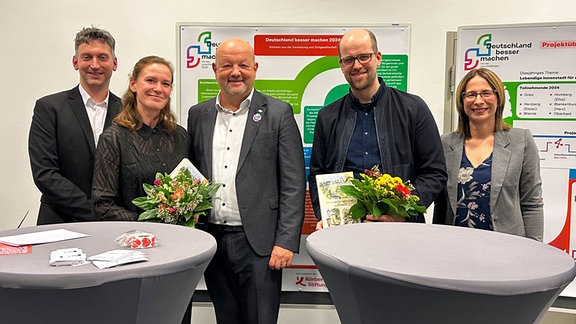 Zwei Frauen und drei Männer halten in formeller Umgebung Blumensträuße in der Hand