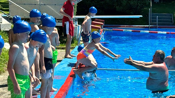 Kinder mit Badekappen stehen am Beckenrand eines Freibades. Ein Mädchen springt ins Wasser.