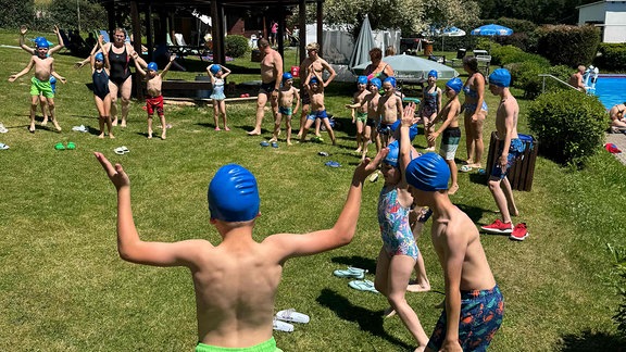 Kinder mit badekappen stehen im Kreis auf einer Wiese und machen Hampfelmänner.