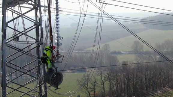 Ein Mensch arbeitet oben auf einem Strommast. Dahinter Wiesen.