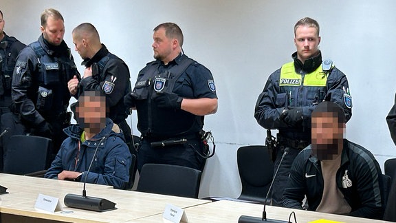 Zwei Männer sitzen an einem Tisch in einem Gerichtssaal. Hinter ihnen stehen mehrere Polizisten.