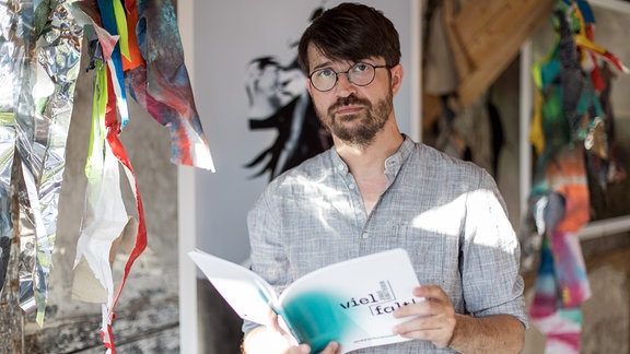 Robert Sorg, ein Mann mit kurzen Haaren, Brille und Bart schaut stehend in die Kamera und hält ein augeschlagenes Buch in den Händen.