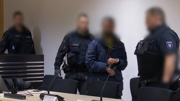 Ein Mann in Handschellen steht neben Polizisten. Auf einem Tisch vor ihm stehen Mikrofone