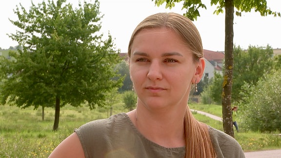 Eine Frau in einem Park. Sie schaut ernst in die Kamera. Sie trägt ein olivgrünes Sommerkleid und hat die Haare zu einem Pferdeschwanz zusammengebunden.