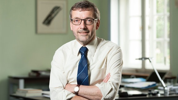 Walter Rosenthal, ein Mann mit Hemd, Schlips und Brille, steht in einem Büro vor einem Schreibtisch.