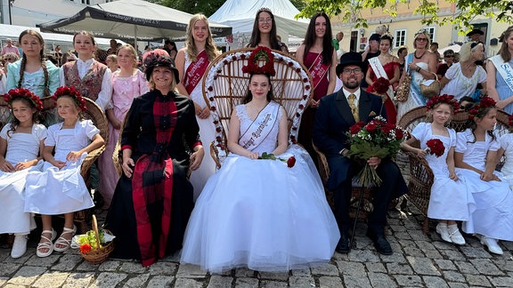 Eine Frau im Kleid mit einer Rosenkrone auf dem Kopf, umgeben von weiteren Personen.