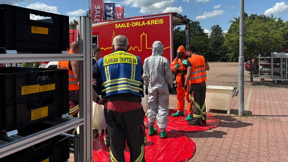 Auf dem Parkplatz eines Baumarkts ziehen Feuerwehrleute Schutzanzüge an.