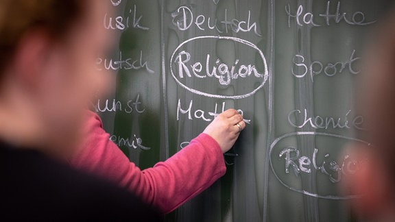 Eine Lehrerin schreibt mit Kreide einen Stundenplan mit Religionsunterricht an eine Tafel.