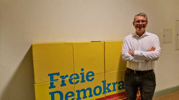 Der FDP-Politiker Robert-Martin Montag steht vor einer Wand und vor gelben großen Würfeln mit dem Schriftzug "Freie Demokraten"