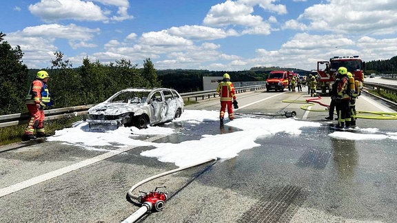 Ein ausgebrannter Pkw auf einer Autobahn, daneben und dahinter Feuerwehrleute