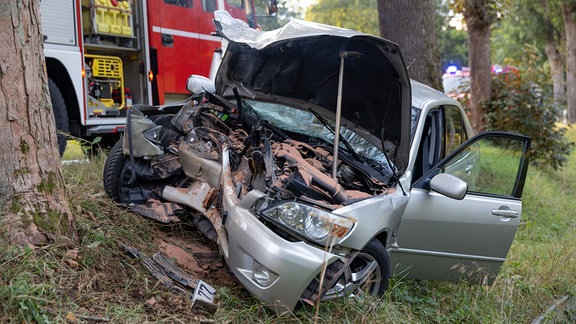 Ein Auto liegt nach einem Unfall mit zerstörter Motorhaube an einem Baum, im Hintergrund ist ein Feuerwehrauto zu sehen.