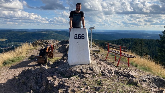 Philipp vom Ruppberg e.V. in Thüringen: Ein Mann steht auf einer Höhenmarkierung auf einem Berg über ihm blauer Himmel mit weißen Wolken, darunter weiter Blick über die Landschaft mit Hügeln und Wäldern