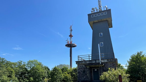 Ein Turm steht neben einem Funkmast auf einer Wiese, auf der Holzbänke stehehn. 