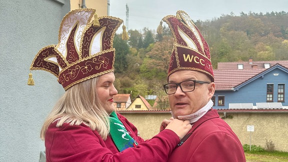 Eine Frau richtet die Krawatte eines Mannes. Beide tragen weinrote Karnevaloutfits.