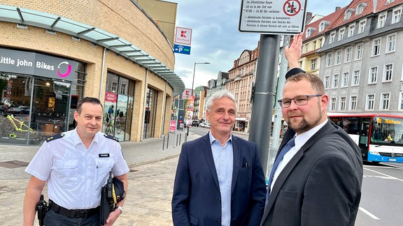 Dei Männer stehen neben einem Ampfelpfleiler, an dem ein Schil einer Alkoholverbotszone hängt. Ein Mann zeigt auf das Schild.