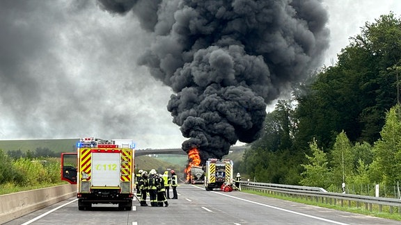 Im Vordergrund ist ein Löschfahrzeug mit Einsatzkräften. Im Hintergrund ein Brennender Tanklastwagen auf der Autobahn.