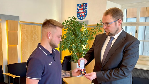 Ein Mann im Anzug überreicht einem jungen Mann eine Urkunde und eine Medaille.