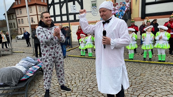 Waltershäuser Bürgermeister Leon Graupner im Schlafanzug und Karnevalspräsident Mario Seifert treten auf einem Markplatz gegeneinander an, Seifert hält ein Mikrofon in der Hand und hält die Siegesfaust hoch, Graupner klatscht strahlend